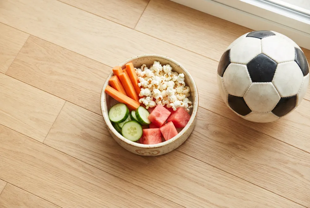 A pet snack bowl of carrot sticks, plain popcorn, cucumber slices and watermelon cubes on a wooden floor next to a soccer ball